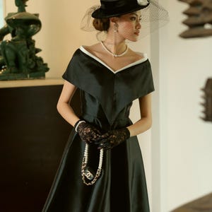 May include: A black satin dress with a white trim neckline, paired with a black hat with a veil. The outfit includes a pearl necklace, black lace gloves, and a pearl and chain handbag. The text "KASHY KATY" is visible at the bottom.