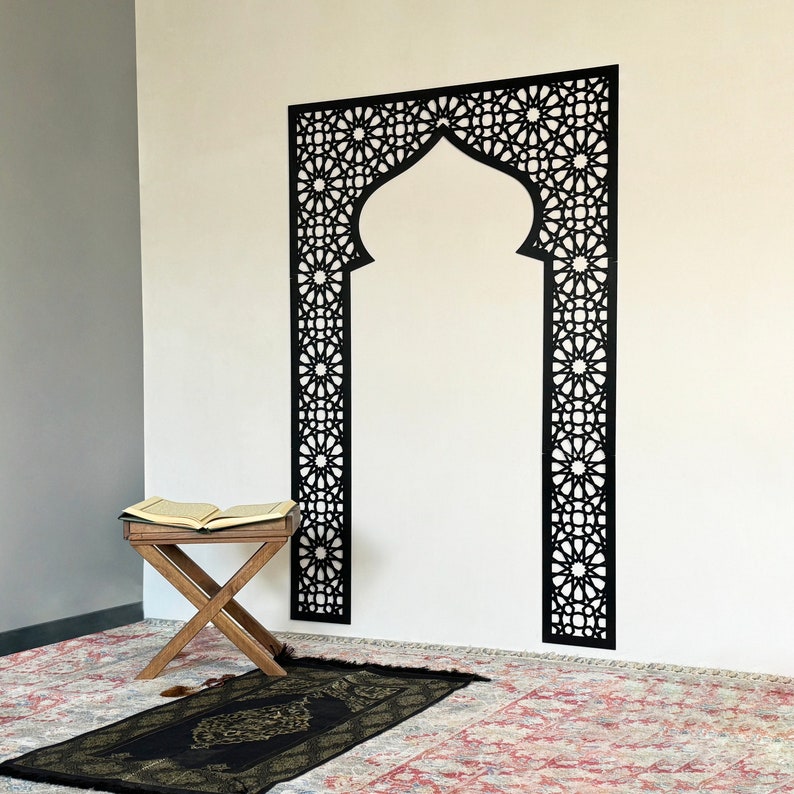 May include: A black decorative wall decal with an intricate geometric design, resembling an archway. A prayer rug and a small wooden stool with an open book are in the foreground.