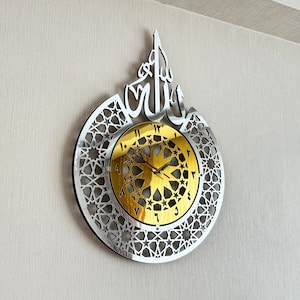 May include: A silver and gold wall clock with an intricate geometric design and Arabic numerals. The clock features the word "Allah" in Arabic script.