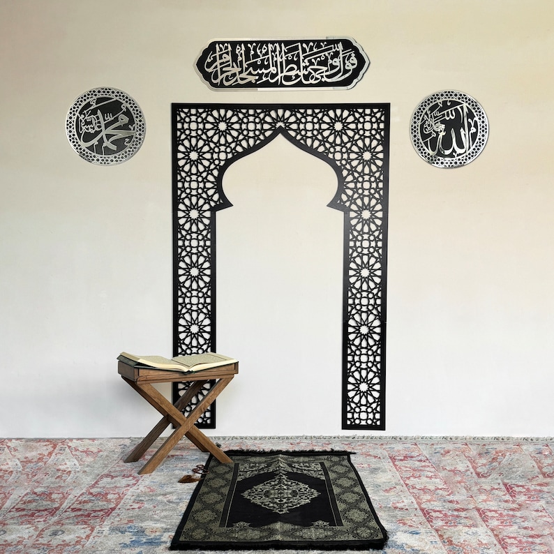 May include: A black prayer rug with a geometric pattern and a black wooden prayer stand with a book on top. The prayer rug is on a patterned rug. There are three silver wall decorations with Arabic calligraphy.