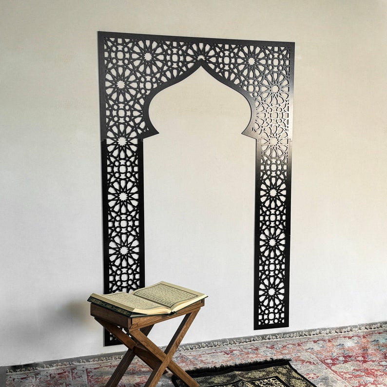 May include: A black metal decorative archway with intricate geometric designs, a wooden prayer stool, and an open book on top of the stool. The archway is mounted on a white wall.