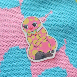 以下が含まれることがあります： 蝶ネクタイと水玉模様のピンクと黄色の漫画キャラクター。キャラクターは足を組んで座っており、キラキラした質感です。