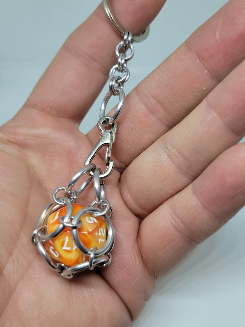 Op de afbeelding: Een zilverkleurige sleutelhanger met een grote, oranje en gele D12 dobbelsteen. De dobbelsteen zit in een kooi-achtige structuur. De sleutelhanger heeft een karabijnhaak en een ring om aan sleutels of tassen te bevestigen.