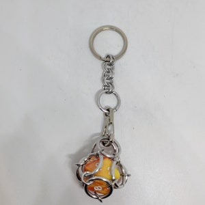 Op de afbeelding: Een sleutelhanger met een zilveren metalen ketting en ring. De sleutelhanger heeft een gele en oranje dobbelsteen met witte cijfers, ingesloten in een zilveren draadkooi. De dobbelsteen is ongeveer 2,5 cm groot.
