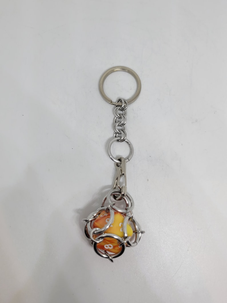 Op de afbeelding: Een sleutelhanger met een zilveren metalen ketting en ring. De sleutelhanger heeft een gele en oranje dobbelsteen met witte cijfers, ingesloten in een zilveren draadkooi. De dobbelsteen is ongeveer 2,5 cm groot.