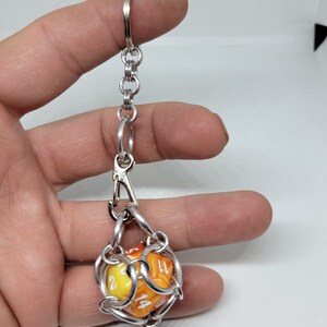 Op de afbeelding: Een zilverkleurige metalen sleutelhanger met een ketting en sluiting, met een D20-dobbelsteen. De dobbelsteen is geel en oranje met witte cijfers. De sleutelhanger is ontworpen om de dobbelsteen vast te houden en weer te geven.