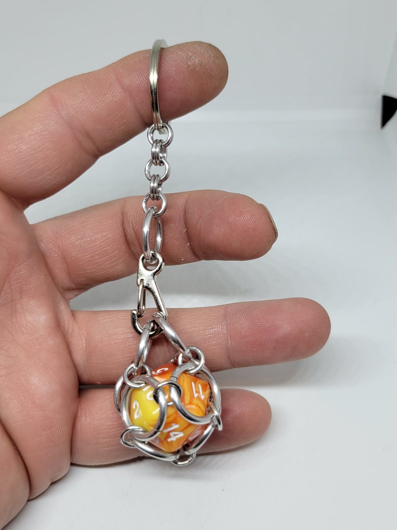 Op de afbeelding: Een zilverkleurige metalen sleutelhanger met een ketting en sluiting, met een D20-dobbelsteen. De dobbelsteen is geel en oranje met witte cijfers. De sleutelhanger is ontworpen om de dobbelsteen vast te houden en weer te geven.