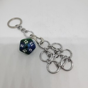Op de afbeelding: Een zilverkleurige sleutelhanger met een blauw-groene D20 dobbelsteen, met witte cijfers. De sleutelhanger heeft ook een chainmail-stijl bedel. Inclusief een splitring en een karabijnhaak voor bevestiging.