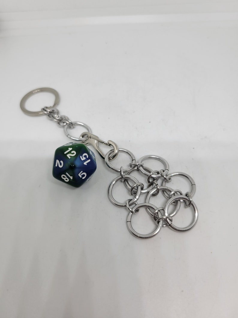 Op de afbeelding: Een zilverkleurige sleutelhanger met een blauw-groene D20 dobbelsteen, met witte cijfers. De sleutelhanger heeft ook een chainmail-stijl bedel. Inclusief een splitring en een karabijnhaak voor bevestiging.