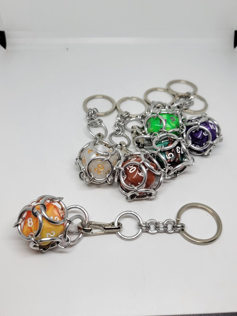 Op de afbeelding: Sleutelhangers met kleurrijke dobbelstenen in zilverkleurige metalen kooien. De dobbelstenen hebben verschillende kleuren, waaronder oranje, groen en paars, met zichtbare cijfers. Elke sleutelhanger heeft een ring om aan sleutels of tassen te bevestigen. De dobbelstenen zijn ongeveer 2,5 cm in diameter.