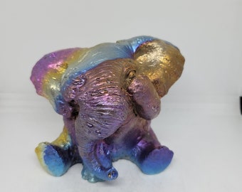Op de afbeelding: Een klein, regenboogkleurig keramiek olifantbeeldje met iriserende kleuren. De olifant zit met zijn slurf omhoog en zijn poten gekruist.
