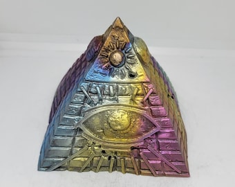 Op de afbeelding: Een veelkleurige piramide met een alziend oogontwerp. De piramide is gemaakt van metaal en heeft een regenboogkleurige, iriserende afwerking.