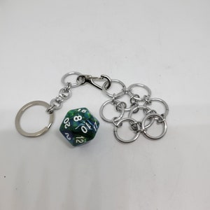 Op de afbeelding: Een zilverkleurige sleutelhanger met een karabijnhaak en een ring, verbonden met een 20-zijdige dobbelsteen. De dobbelsteen is groen en blauw met witte cijfers. Een zilveren accessoire in kettingstijl is ook bevestigd.