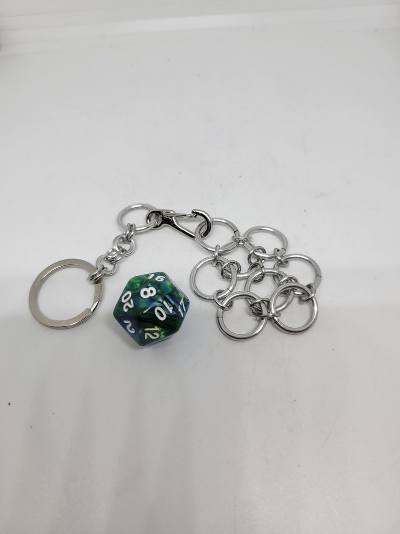 Op de afbeelding: Een zilverkleurige sleutelhanger met een karabijnhaak en een ring, verbonden met een 20-zijdige dobbelsteen. De dobbelsteen is groen en blauw met witte cijfers. Een zilveren accessoire in kettingstijl is ook bevestigd.