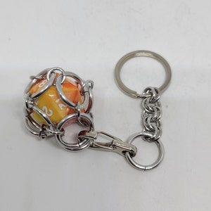 Op de afbeelding: Een sleutelhanger met een D20-dobbelsteen in een zilveren kettingserie-ontwerp. De dobbelsteen is oranje en geel met witte cijfers. De sleutelhanger heeft een karabijnhaak en een sleutelring.
