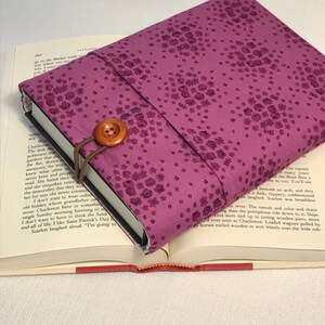 May include: A purple book cover with a floral pattern and a brown button closure. The cover is made of fabric and has a soft, textured feel.