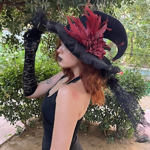 May include: A black witch hat with a wide brim, adorned with a large burgundy flower and feathers. The hat has a curved, pointed tip. Black lace gloves and a black dress complete the gothic ensemble, set against a backdrop of green foliage.