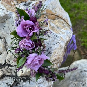 May include: A floral crown featuring three lavender-colored roses, green leaves, and small purple flowers. The crown is adorned with a delicate wire base and purple ribbon ties, perfect for a wedding or special occasion.