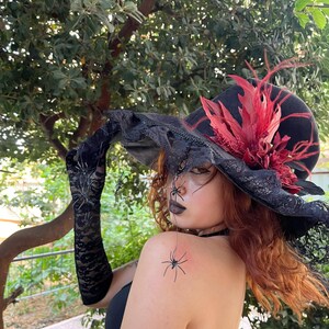 May include: A black witch hat with a wide brim, adorned with red feathers and lace trim. The hat is part of a Halloween costume, with black lace gloves and spider decorations.