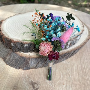 May include: A boutonniere featuring a vibrant arrangement of dried flowers including bright orange, turquoise, and purple sprigs, a soft pink bunny tail grass, and two large pink and deep red strawflowers, all bound together with twine.