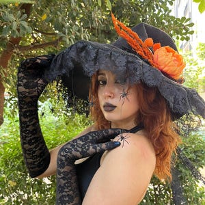 May include: A black witch hat with a wide brim, trimmed with black lace and adorned with orange and peach-colored floral accents. The hat is paired with long, black lace gloves. Small spider decorations are visible.
