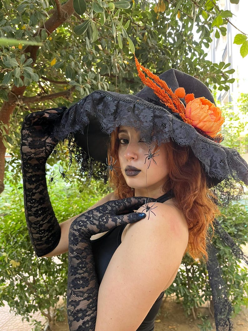 May include: A black witch hat with a wide brim, trimmed with black lace and adorned with orange and peach-colored floral accents. The hat is paired with long, black lace gloves. Small spider decorations are visible.