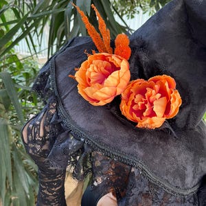 May include: A black witch hat with orange floral accents and black lace trim. The hat is decorated with two large orange flowers and several smaller orange decorative elements. The brim is trimmed with black lace.