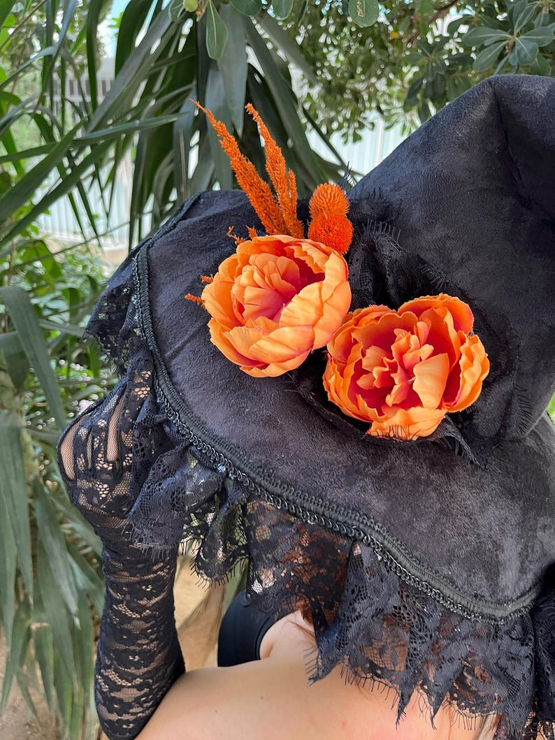 May include: A black witch hat with orange floral accents and black lace trim. The hat is decorated with two large orange flowers and several smaller orange decorative elements. The brim is trimmed with black lace.