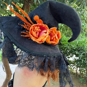 May include: Black witch hat with a wide brim, adorned with orange and pink flowers and sprigs. The hat features a lace trim and is perfect for Halloween or costume parties.