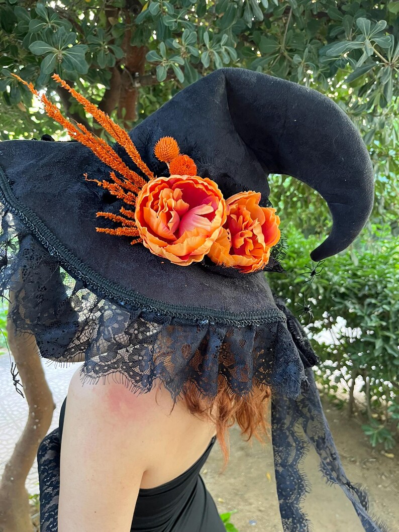 May include: Black witch hat with a wide brim, adorned with orange and pink flowers and sprigs. The hat features a lace trim and is perfect for Halloween or costume parties.