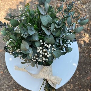Pode incluir: Um bouquet de folhas de eucalipto secas, pequenas flores brancas de gips&oacute;fila e folhagem verde. Os caules est&atilde;o reunidos e atados com uma fita tecida de cor creme com textura, que apresenta um pequeno padr&atilde;o de losangos repetido.