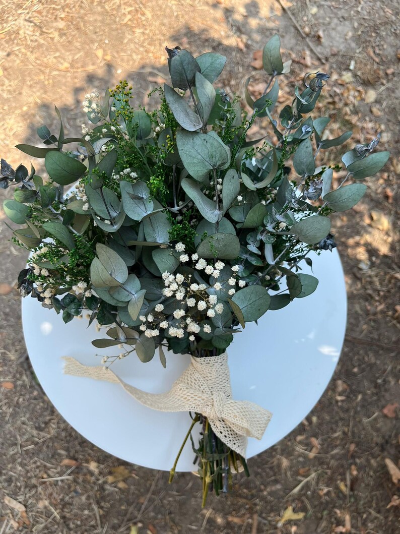 Pode incluir: Um bouquet de folhas de eucalipto secas, pequenas flores brancas de gips&oacute;fila e folhagem verde. Os caules est&atilde;o reunidos e atados com uma fita tecida de cor creme com textura, que apresenta um pequeno padr&atilde;o de losangos repetido.