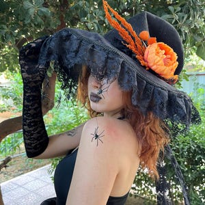 May include: Black witch hat with a wide brim, adorned with black lace trim and a decorative orange flower and accents. The hat is part of a Halloween costume, with black lace gloves and spider decorations.