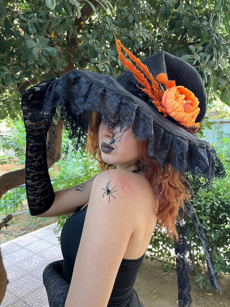 May include: Black witch hat with a wide brim, adorned with black lace trim and a decorative orange flower and accents. The hat is part of a Halloween costume, with black lace gloves and spider decorations.