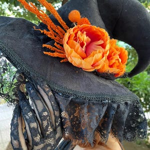 May include: A black witch hat with a wide brim, adorned with orange flowers and sprigs. The hat features a lace trim and is decorated with a large orange flower. The hat is a Halloween costume accessory.
