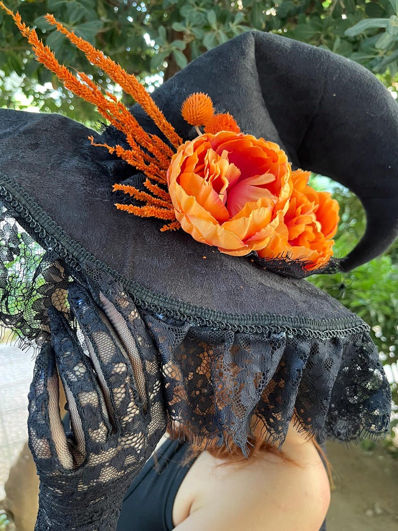 May include: A black witch hat with a wide brim, adorned with orange flowers and sprigs. The hat features a lace trim and is decorated with a large orange flower. The hat is a Halloween costume accessory.