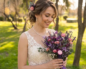 Pode incluir: Um vestido branco de renda sem mangas com decote em V, um colar de pérolas e brincos pendentes. Um acessório de cabelo e um buquê de flores secas em tons de rosa, roxo e verde, atado com uma fita roxa, segurado à frente do corpo.