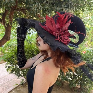 May include: A black witch hat with a wide brim, adorned with red feathers and a large, dark red flower. The hat has a curved shape and is trimmed with black lace. The image shows a person wearing the hat, along with black lace gloves.