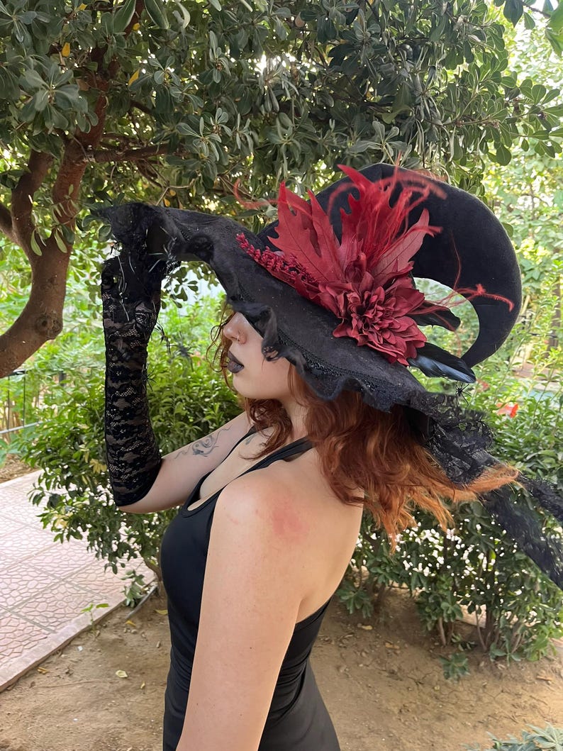 May include: A black witch hat with a wide brim, adorned with red feathers and a large, dark red flower. The hat has a curved shape and is trimmed with black lace. The image shows a person wearing the hat, along with black lace gloves.