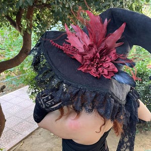 May include: A black witch hat with a wide brim, adorned with a large burgundy flower and feathers. The hat features a black lace trim around the brim. The hat is part of a costume.