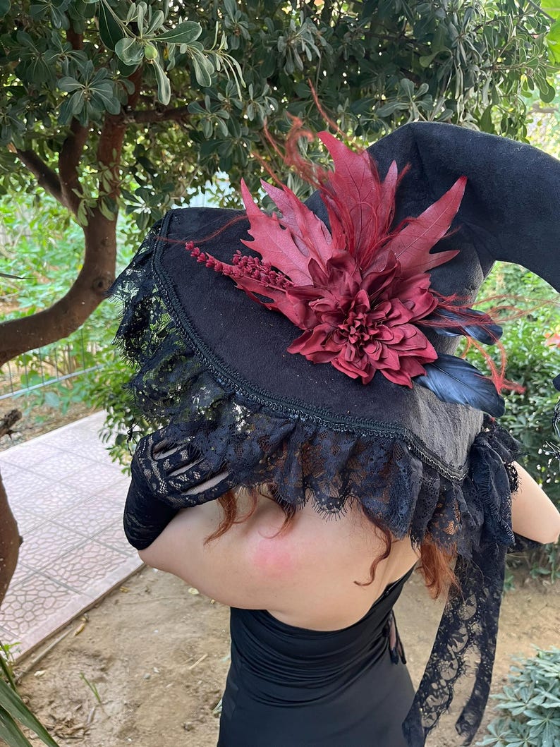 May include: A black witch hat with a wide brim, adorned with a large burgundy flower and feathers. The hat features a black lace trim around the brim. The hat is part of a costume.