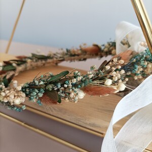 A dried flower crown with white, beige, and teal flowers. The crown is on a wooden surface with a white ribbon.