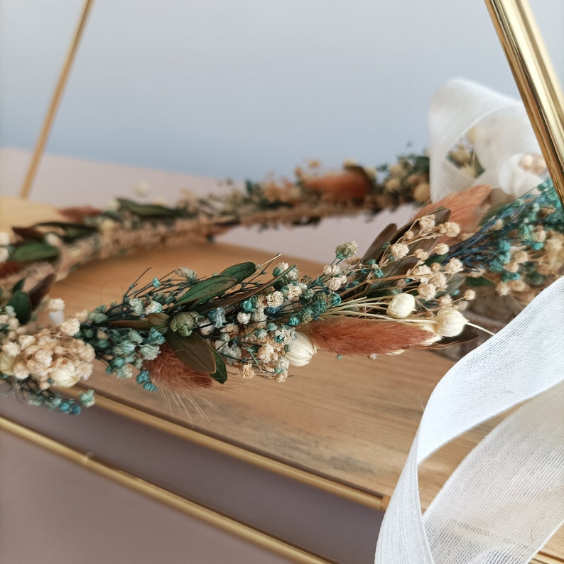 A dried flower crown with white, beige, and teal flowers. The crown is on a wooden surface with a white ribbon.