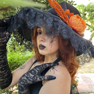 May include: A black witch hat with a wide brim, adorned with black lace, orange flowers, and decorative spiders. The hat is part of a Halloween costume, paired with black lace gloves.