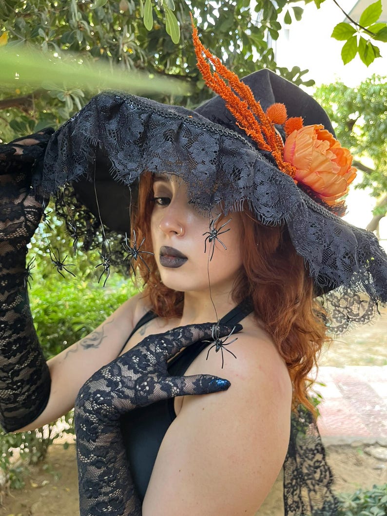 May include: A black witch hat with a wide brim, adorned with black lace, orange flowers, and decorative spiders. The hat is part of a Halloween costume, paired with black lace gloves.