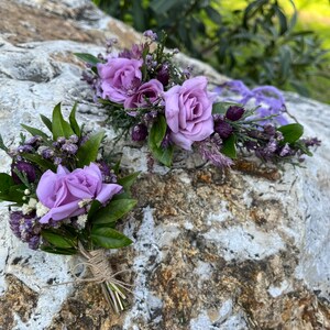 May include: Two floral arrangements featuring lavender roses, green leaves, and purple accents. One arrangement is a boutonniere tied with twine, and the other is a larger floral piece, possibly a headpiece or corsage. The flowers rest on a textured stone surface.