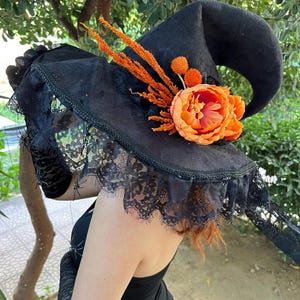May include: Black witch hat with a curved brim, adorned with orange floral accents and a large orange flower. The hat features a black lace veil and is perfect for Halloween or costume parties.
