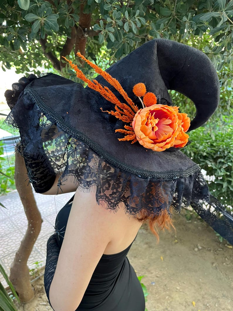 May include: Black witch hat with a curved brim, adorned with orange floral accents and a large orange flower. The hat features a black lace veil and is perfect for Halloween or costume parties.