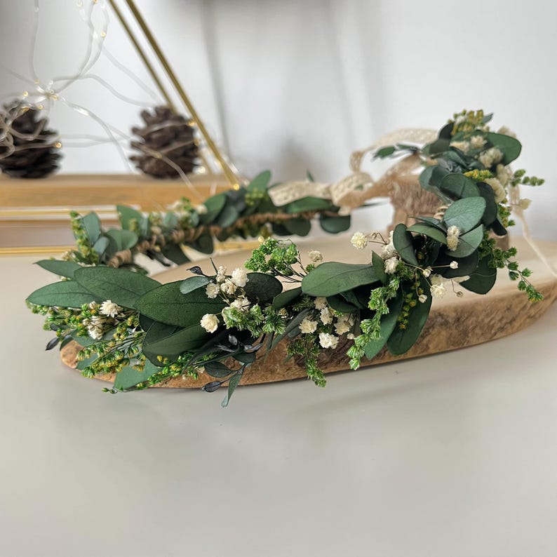 Pode incluir: Uma coroa de flores com folhas de eucalipto verde-escuras, pequenas flores brancas de gips&oacute;fila e folhagem verde delicada. O acess&oacute;rio circular &eacute; constru&iacute;do sobre uma base fina e flex&iacute;vel e inclui uma fita de cor bege claro.