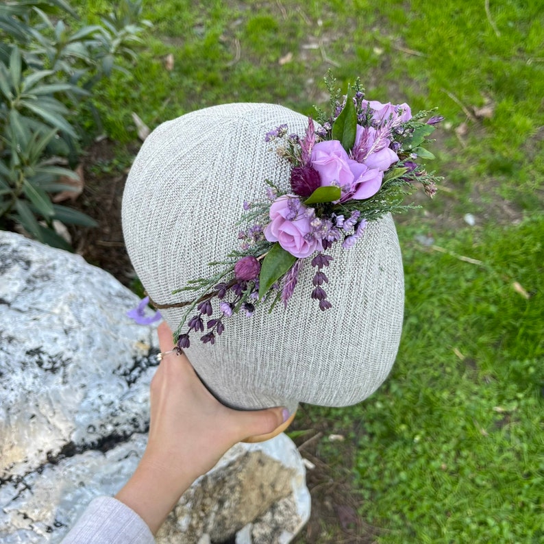 May include: A light gray fabric headpiece adorned with a floral arrangement. The arrangement features lavender-colored roses, sprigs of purple flowers, and green leaves. The headpiece is held by a hand, with a natural background of grass and stone.